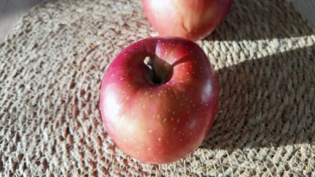 Two fresh organic red apples with stems resting on woven placemat under warm natural sunlight.