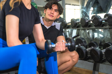 Fototapeta premium Handsome Asian male personal trainer helping and instructing a fit sportswoman during workout with dumbbells exercise equipment in modern gym