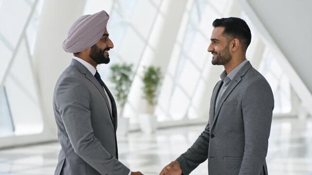 Two professional businessmen in grey suits, one wearing a turban, smile as they engage in a firm handshake in a bright, modern corporate lobby, sealing a deal or greeting each other.