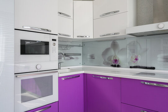 compact corner kitchen combines glossy purple lower cabinets with crisp white upper units and built‑in appliances, accentuated by a floral glass splashback for a bold, modern look