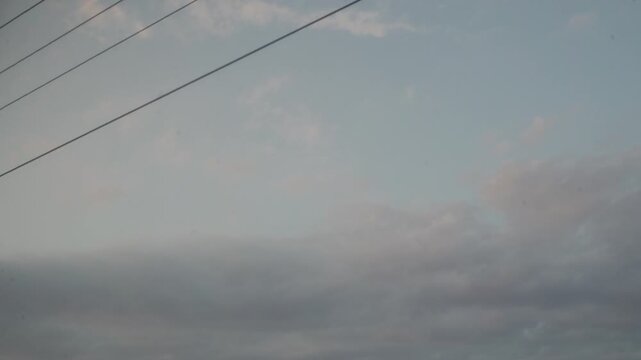 Delicate clouds drift quietly across a vast, overcast sky. Thin power lines stretch overhead, evoking a calm and nostalgic feeling during a long journey.