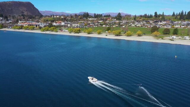 Aerial view of motorboat with person on wakeboard riding on lake wanaka with scenic town and mountains in background on sunny day.