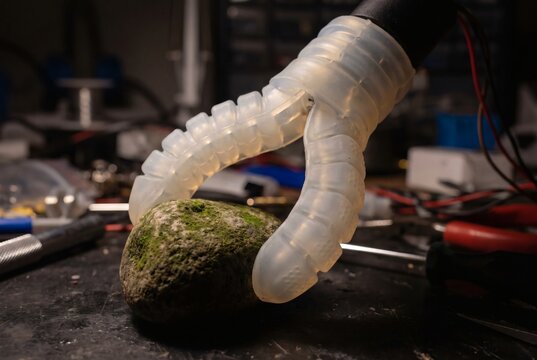 Transparent silicone soft robotic gripper holding mossy stone on laboratory workbench with tools, soft robotics and flexible automation concept