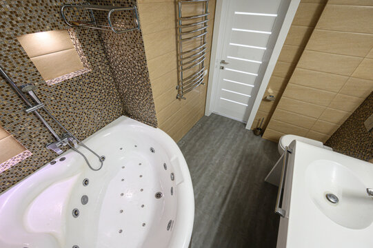 compact bathroom features a white corner jacuzzi tub with jets, mosaic-tiled shower walls, a chrome towel radiator, beige tiles, and a white door with horizontal slats