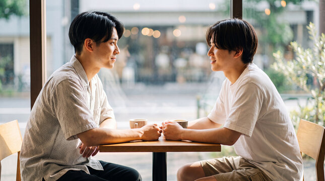 カフェのテーブルで手を置き見つめ合う二人の若い男性 ｜ 自然光