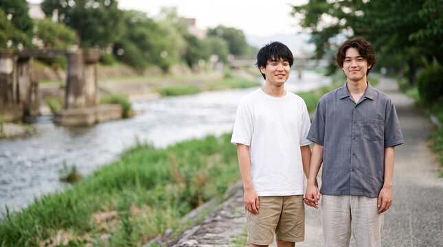 夏の川沿いの道で手を繋ぎ優しく微笑み合う男性二人 ｜ 爽やかな風