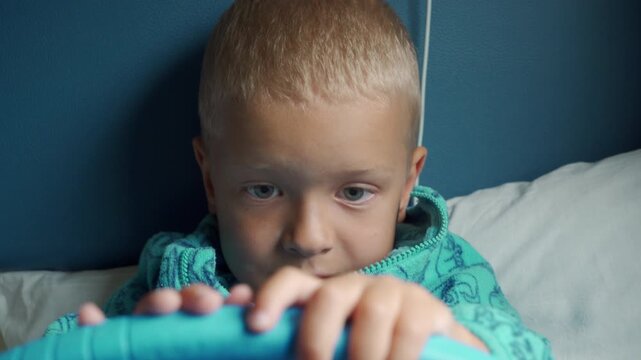 A focused young boy plays with a tablet while resting in a cozy bed. His curious expression fills the quiet space with gentle warmth and calm.