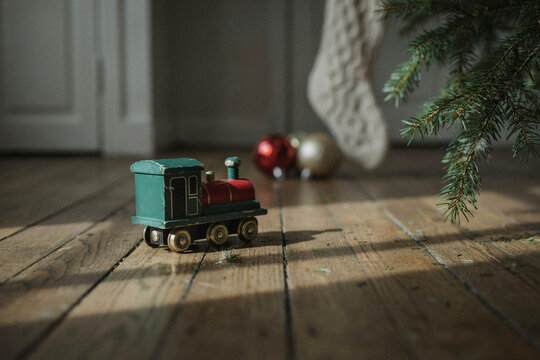Small toy train on rustic wooden floor near Christmas tree, cozy holiday home interior with soft lights and ornaments for seasonal decor