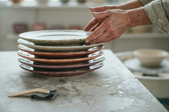 Stack of ceramic plates in neutral tones on kitchen counter, close-up hands arranging stoneware tableware for home dining and food styling