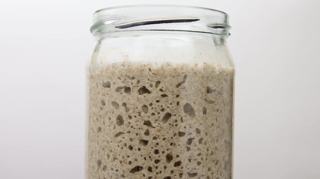 Active sourdough starter with air bubbles fermenting in glass jar on white background.