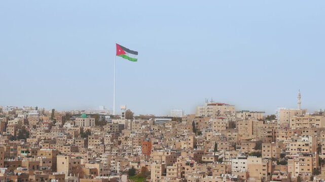 Large Jordan flag waving on a big pole above Amman - the capital city. Many buildings below