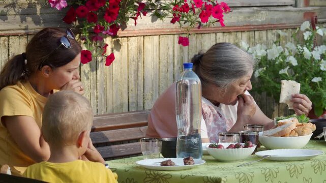 A happy family enjoys an outdoor lunch together at a table. They are eating and chatting warmly in a cozy, rustic garden setting surrounded by bright flowers.