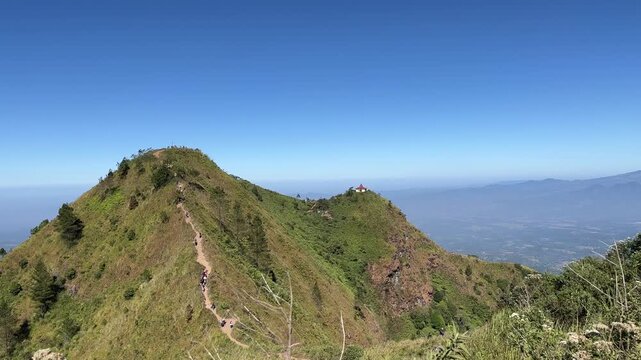Panoramic view of Mount Andong peak with hikers along the grassy ridge trail under a clear blue sky, showing surrounding valleys and lush green slopes.