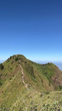 Panoramic view of Mount Andong peak with hikers along the grassy ridge trail under a clear blue sky, showing surrounding valleys and lush green slopes.