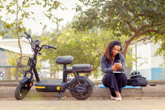 Young Black Brazilian woman checking her smartphone during a break from her e-bike commute, representing sustainable mobility, connected urban lifestyle, and eco-friendly transportation.