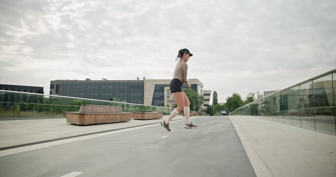 A young beautiful girl in sportswear and a cap does exercises with a skipping rope and plays sports in an urban setting on a bridge with a pedestrian area and benches against the backdrop of new concr