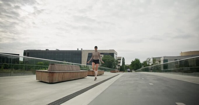 A young beautiful girl in sportswear and a cap does exercises with a skipping rope and plays sports in an urban setting on a bridge with a pedestrian area and benches against the backdrop of new concr
