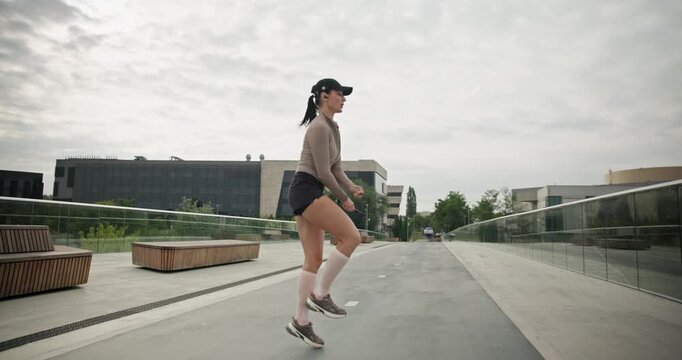 A young beautiful girl in sportswear and a cap does exercises with a skipping rope and plays sports in an urban setting on a bridge with a pedestrian area and benches against the backdrop of new concr