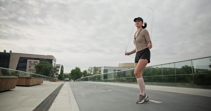 A young beautiful girl in sportswear and a cap does exercises with a skipping rope and plays sports in an urban setting on a bridge with a pedestrian area and benches against the backdrop of new concr