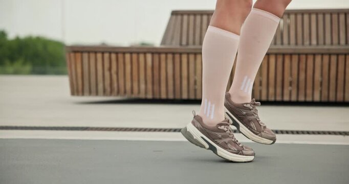 A young beautiful girl in sportswear and a cap does exercises with a skipping rope and plays sports in an urban setting on a bridge with a pedestrian area and benches against the backdrop of new concr