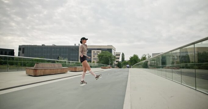 A young beautiful girl in sportswear and a cap does exercises with a skipping rope and plays sports in an urban setting on a bridge with a pedestrian area and benches against the backdrop of new concr