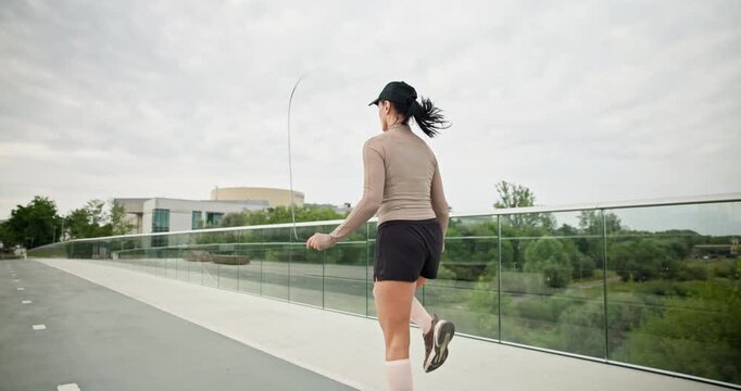 A young beautiful girl in sportswear and a cap does exercises with a skipping rope and plays sports in an urban setting on a bridge with a pedestrian area and benches against the backdrop of new concr