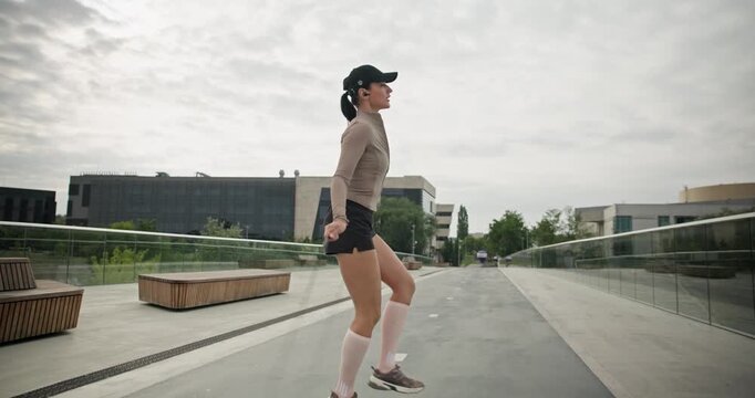 A young beautiful girl in sportswear and a cap does exercises with a skipping rope and plays sports in an urban setting on a bridge with a pedestrian area and benches against the backdrop of new concr