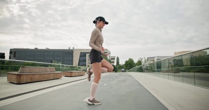 A young beautiful girl in sportswear and a cap does exercises with a skipping rope and plays sports in an urban setting on a bridge with a pedestrian area and benches against the backdrop of new concr