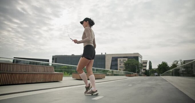 A young beautiful girl in sportswear and a cap does exercises with a skipping rope and plays sports in an urban setting on a bridge with a pedestrian area and benches against the backdrop of new concr