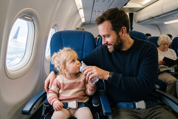 Fototapeta premium A man sits next to a young child on an airplane. He holds a water bottle to help her drink. The child looks up at him with interest. In the background, an older woman reads a book.