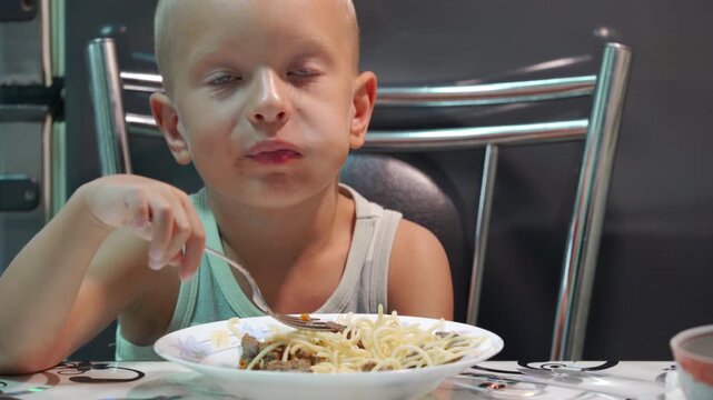 A funny little boy happily eats a delicious plate of spaghetti. He sits contentedly at a kitchen table while enjoying his favorite meal.