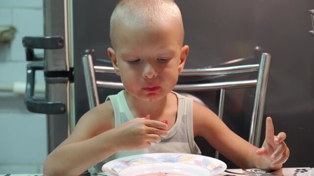 A content little boy focuses on eating a piece of juicy watermelon. He sits happily at a kitchen table with a messy face, fully enjoying his sweet summer snack.