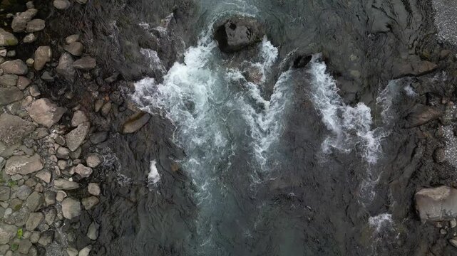 A rushing river flows over rocks creating white water currents amidst smooth stones along its banks, capturing an aerial perspective on nature's raw power within a serene yet dynamic landscape.