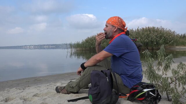 Traveler resting on a quiet lakeshore while eating a sandwich during a cycling adventure. Peaceful wide shot with calm water, reeds, backpack, bicycle helmet, and relaxing outdoor atmosphere