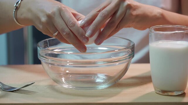 Gentle hands carefully crack a fresh egg into a clear glass bowl. She prepares a comforting breakfast at a wooden kitchen table, creating a warm, cozy feeling of home.