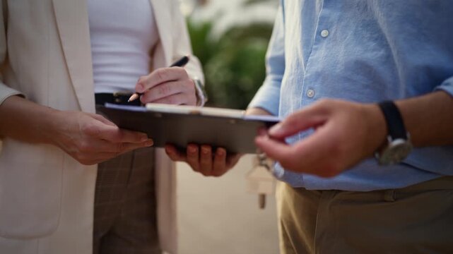 Close-up of homebuyer signing real estate purchase contract on clipboard before receiving house keys from real estate agent, successfully completing property transaction and becoming new homeowner