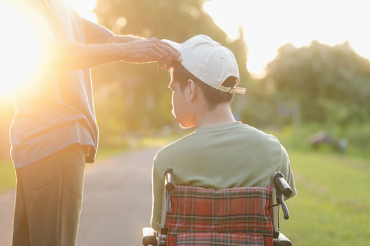 Side view of a male caregiver or parent adjusting a cap and comforting a young man in a wheelchair during golden hour sunset, showing warm family support, love and caring outdoor lifestyle.