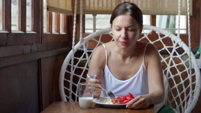 A calm woman peacefully eats her healthy breakfast of eggs and tomatoes at a small table. She sits contentedly inside a cozy wooden veranda in a hanging macrame chair.