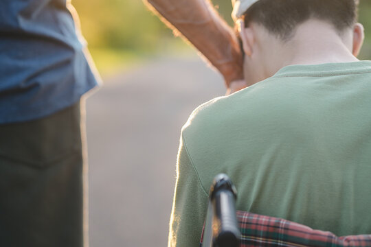 Back view focus on shoulders of a sad young man in a wheelchair receiving comforting hand touch from a caregiver, showing emotional support, empathy and caring human connection.