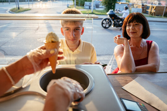 Teenage boy receiving ice cream cone while mature mother waits at sunny street kiosk