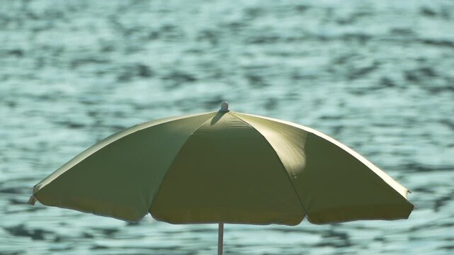 Sun umbrella shade provides relief against a shimmering turquoise ocean surface at a coastal resort. Static eye-level long shot.