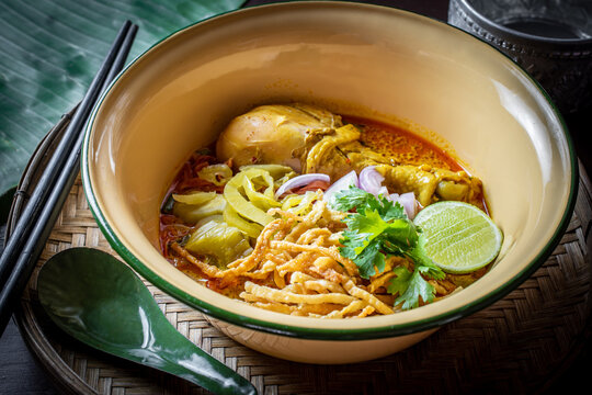 Traditional Northern Thai chicken curry noodle soup, known as Khao Soi, served in a vintage ceramic bowl with crispy egg noodles, fresh lime, pickled mustard greens, and red onion garnish. 