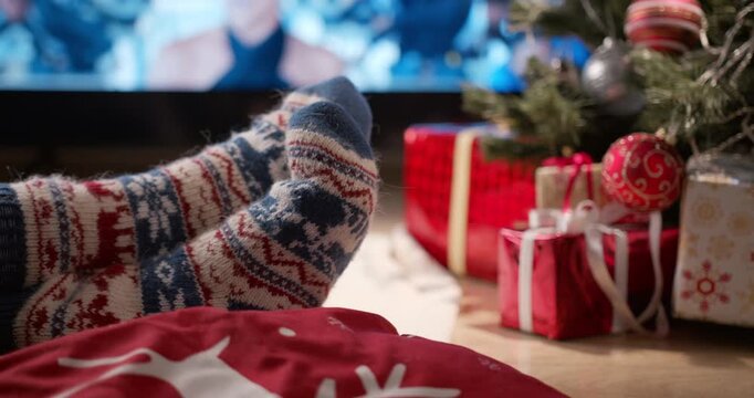 Feet in cozy patterned knitted socks rest near a decorated Christmas tree surrounded by red wrapped gifts and ornaments.