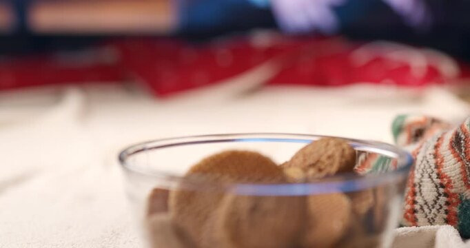 A glass bowl of cookies sits on a soft blanket beside a foot in a cozy patterned Christmas sock in a warm holiday setting.