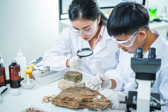 Scientist laboratory rock microscope geology research safety goggle student mineral sample examination during earth science study, focused teamwork in bright classroom workspace