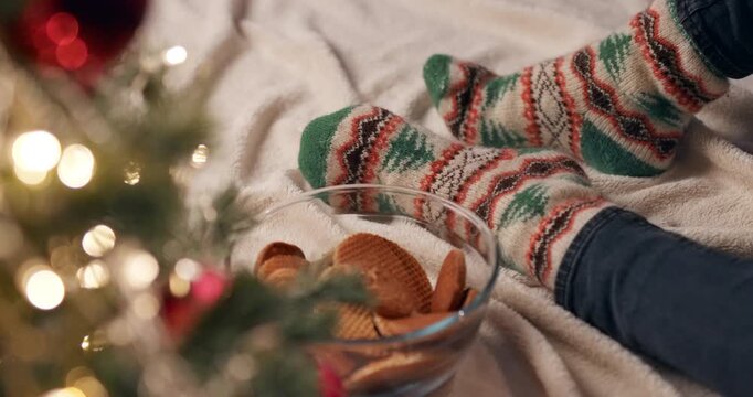 A hand reaches into a bowl of cookies while a person relaxes in patterned knitted socks by a glowing Christmas tree.