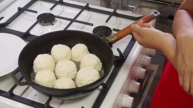 A proud cook gently fries round cottage cheese patties in hot oil using a black pan. She stands happily next to a white stove inside a clean, cozy kitchen.