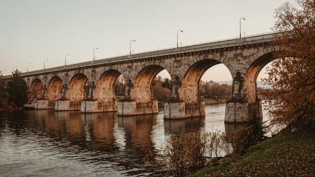 Bridge over river with arches during autumn, with trees showing fall colors and calm water reflecting the structure. Old stone bridge scenery.