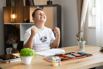 Fototapeta premium Young boy student celebrating achievement with fists clenched and eyes closed in relief after finishing homework at home desk