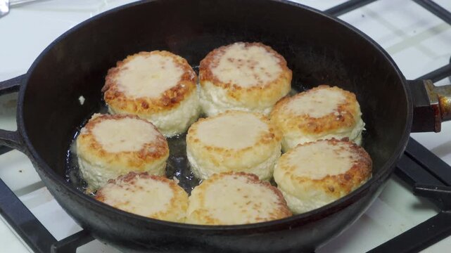 Seven delicious, golden cottage cheese pancakes sizzle eagerly in hot oil. They fry beautifully inside a heavy black cast iron pan on top of a clean white kitchen stove.